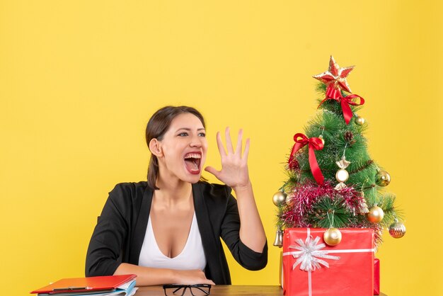 Young woman in suit calling someone near decorated Christmas tree at office on yellow 