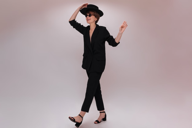 Young woman in suit and hat moving on isolated background. Attractive short-haired lady in black jacket and pants has walk on white backdrop