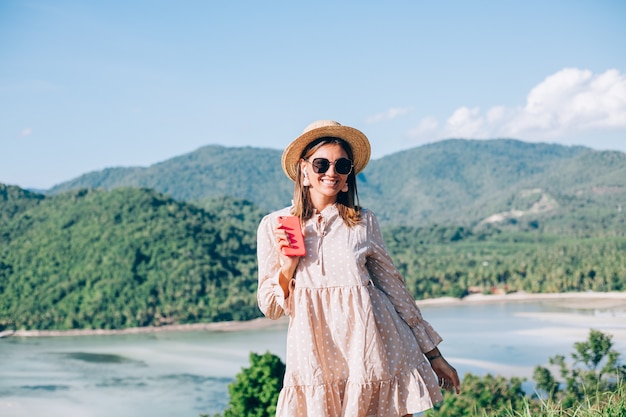 Young woman in summer cute dress, straw hat and sunglasses dancing with smartphone on hand