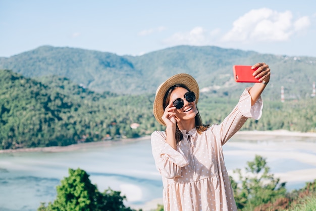 Молодая женщина в летнем милом платье, соломенной шляпе и солнцезащитных очках делает видеозвонок со своего смартфона