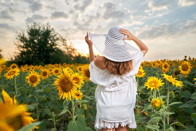 Молодая женщина в летней шляпе стоит на поле с подсолнухами