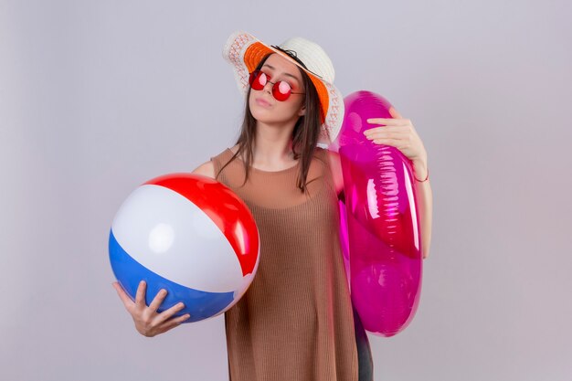 Young woman in summer hat wearing red sunglasses holding inflatable ball and ring looking aside with pensive expression thinking trying make a choice standing over white background