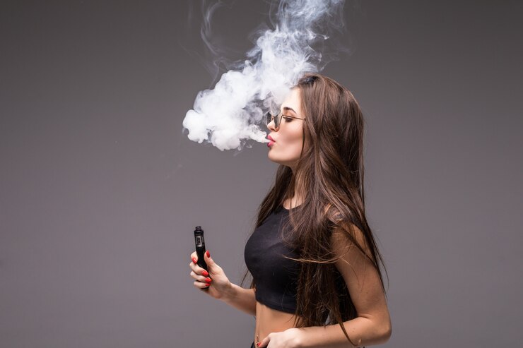 Free Photo young woman in sunglasses and black clothes vaping and blowing smoke of electronic cigarette on gray wall