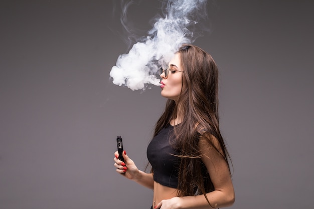 Young woman in sunglasses and black clothes vaping and blowing smoke of electronic cigarette on gray wall