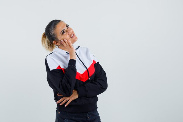 Young woman in sweater and black jeans standing in thinking pose, putting hand on cheek and looking pensive , front view.