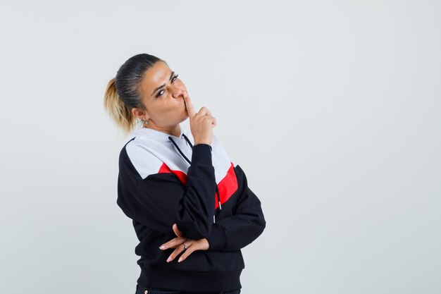 Young woman in sweater and black jeans standing in thinking pose, putting index finger on mouth and looking pensive , front view.