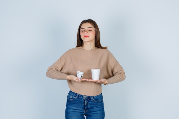 Young woman in sweater, jeans holding paper cups, closing eyes and looking dreamy , front view.