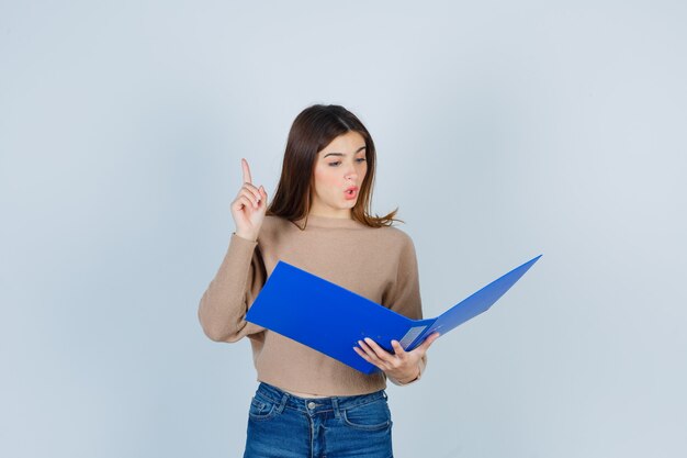Young woman in sweater, jeans looking in folder, pointing up and looking surprised , front view.