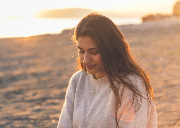 Молодая женщина в свитере сидит на песчаном берегу моря