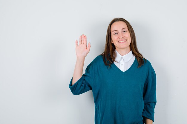 Young woman in sweater over white shirt greeting with open hand and looking merry , front view.