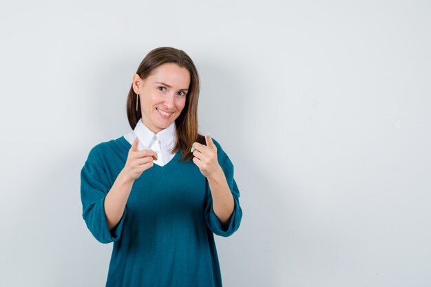 Young woman in sweater over white shirt pointing forward and looking jovial , front view.