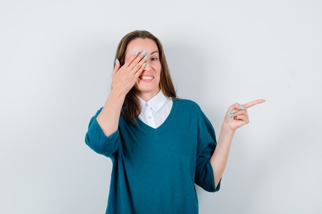 Young woman in sweater over white shirt pointing right, keeping hand on eye and looking clueless , front view.