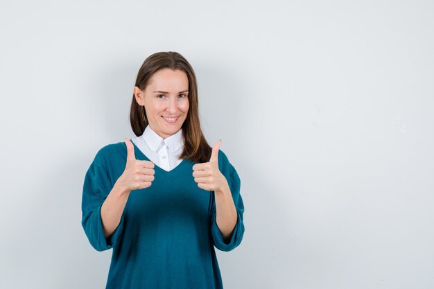 Молодая женщина в свитере над белой рубашкой показывает двойные пальцы вверх и выглядит счастливой, вид спереди.