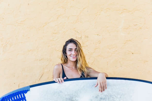 Young woman in swimsuit with surfboard at yellow wall