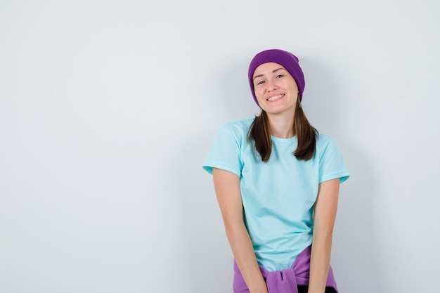 Young woman in t-shirt, beanie posing while standing and looking cheerful , front view.