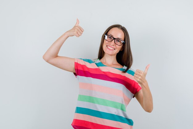 Молодая женщина в футболке, очках показывает палец вверх и выглядит веселой, вид спереди.