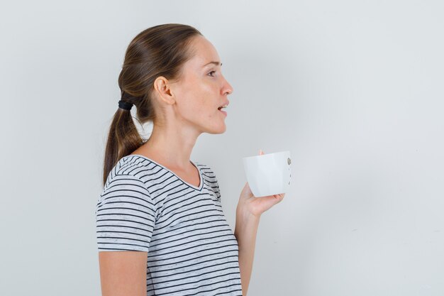 Young woman in t-shirt holding cup of tea and looking focused .