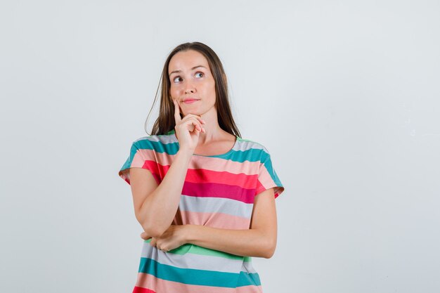 Young woman in t-shirt holding finger on cheek and looking pensive , front view.