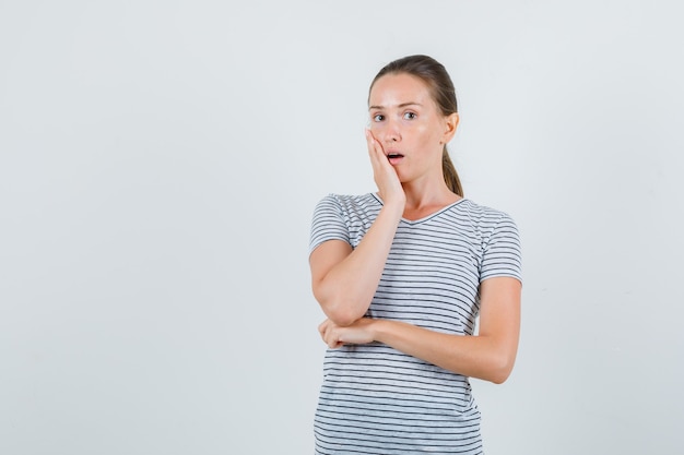 Young woman in t-shirt holding hand on cheek and looking surprised , front view.