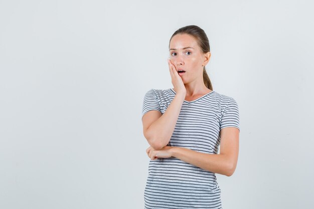 Young woman in t-shirt holding hand on cheek and looking surprised , front view.