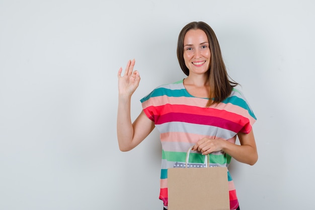 Young woman in t-shirt holding paper bags with ok sign and looking happy , front view.