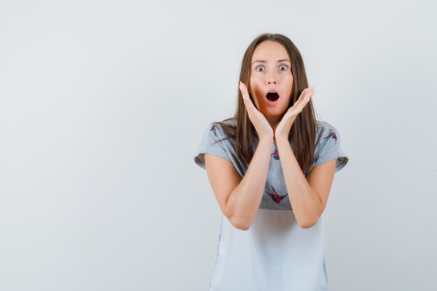 Young woman in t-shirt holding raised hands near mouth and looking surprised , front view.