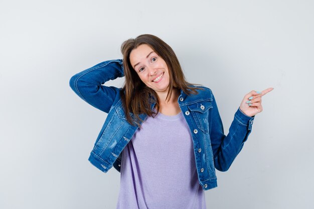 Young woman in t-shirt, jacket with hand behind head, pointing right and looking indecisive , front view.