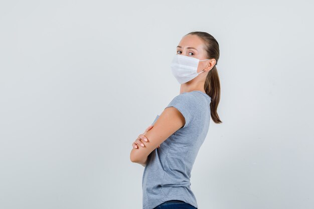 Giovane donna in t-shirt, maschera, jeans in piedi con le braccia incrociate e guardando fiducioso.