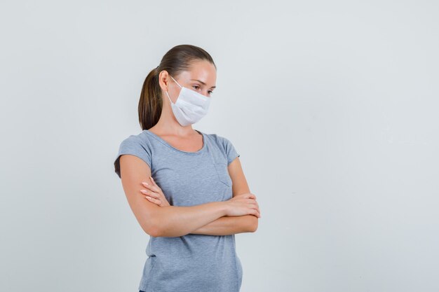 Young woman in t-shirt, mask standing with crossed arms and looking pensive , front view.