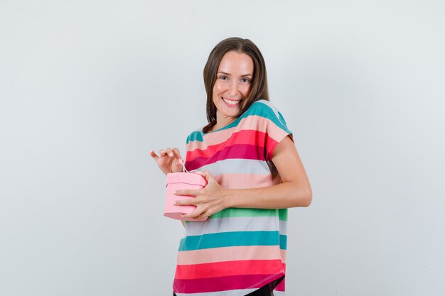 Молодая женщина в футболке, штанах держит подарочную коробку и рад, вид спереди.