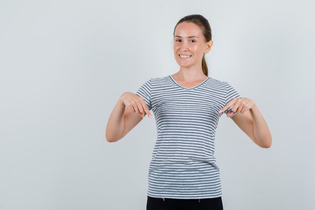 Young woman in t-shirt, pants pointing down and looking cheerful , front view.