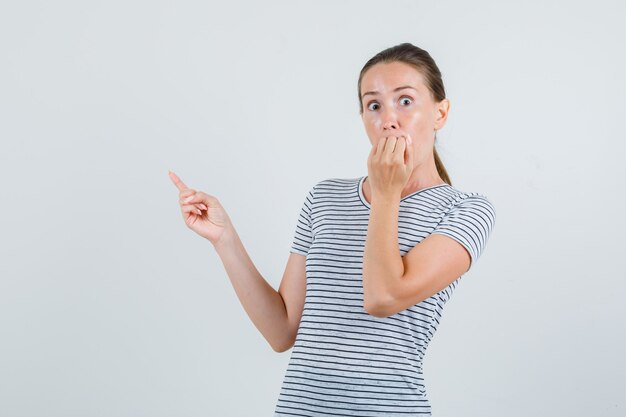 Young woman in t-shirt pointing to side while biting fist and looking scared , front view.