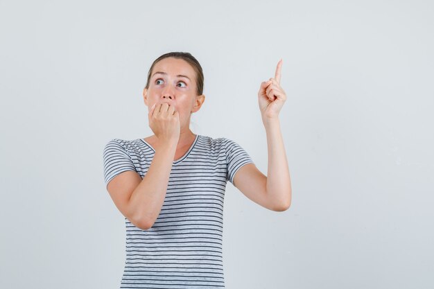 Young woman in t-shirt pointing up while biting fist and looking scared , front view.