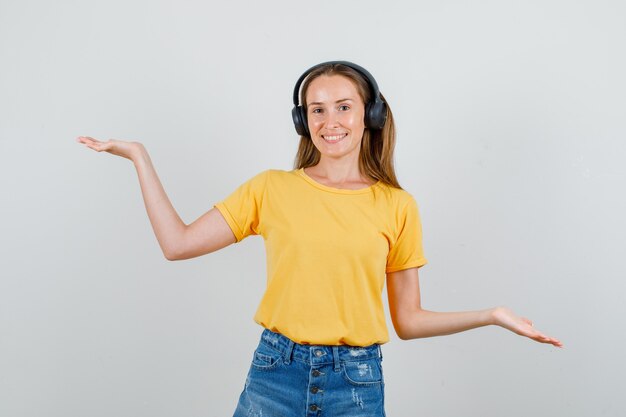 Young woman in t-shirt, shorts, headphones raising hands to show balance and looking glad