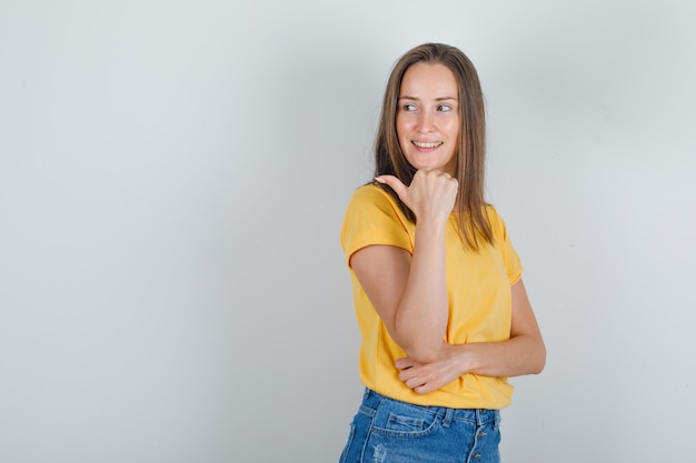 Tシャツを着た若い女性、親指で横を向いて陽気に見えるショートパンツ