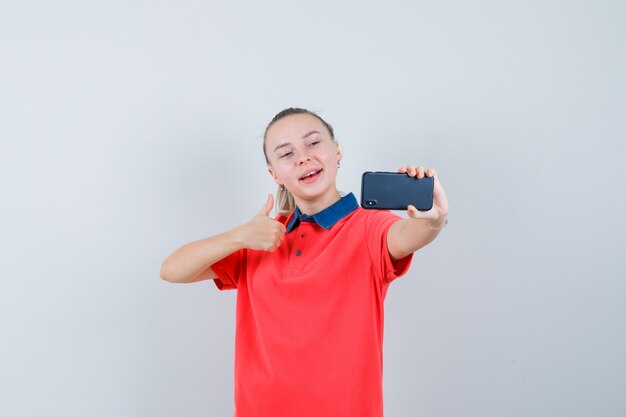 Young woman in t-shirt showing thumb up while taking selfie and looking cheery