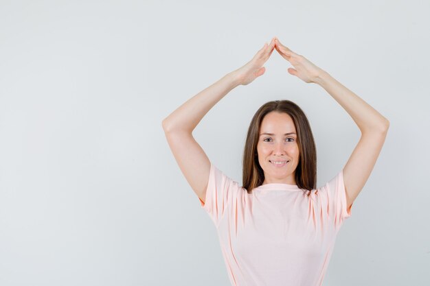 Young woman in t-shirt, skirt gesturing house roof over head and looking cheery , front view.