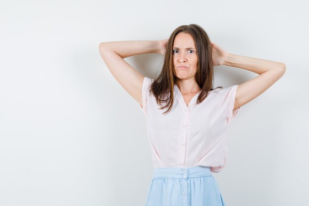 Young woman in t-shirt, skirt holding hands behind head and looking angry , front view.