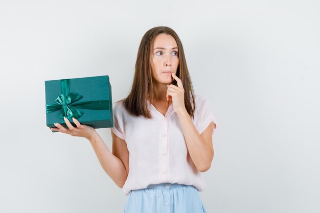 Young woman in t-shirt, skirt holding present box and looking pensive , front view.