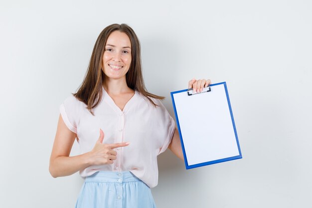 Young woman in t-shirt, skirt pointing at clipboard and looking merry , front view.