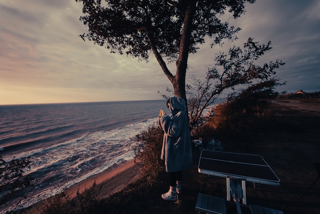 若い女性がスマートフォンで海の夕日の写真を撮る