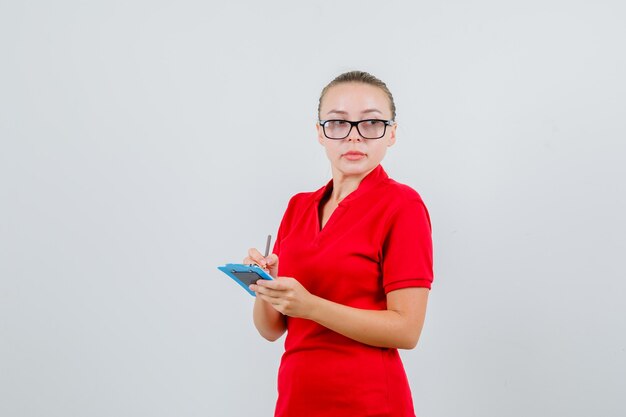赤いTシャツ、メガネ、物思いにふけるクリップボードにメモを取る若い女性