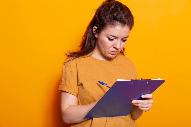 Young woman taking notes on textbook papers with pen in studio. Concentrated person writing on notebook files and signing documents and paperwork about project on notepad clipboard.