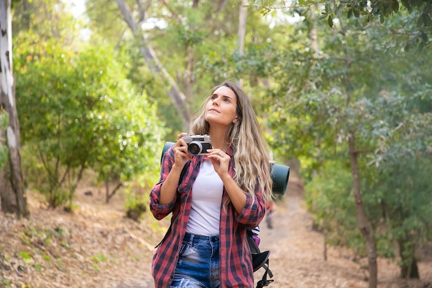 Young woman taking photo of landscape with camera and standing on forest road. Caucasian long-haired female tourist shooting nature in woods. Backpacking tourism, adventure and summer vacation concept