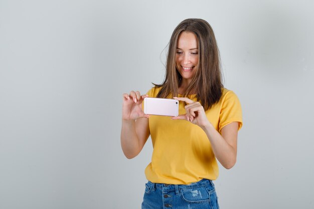 Young woman taking photo on phone in t-shirt, shorts and looking glad