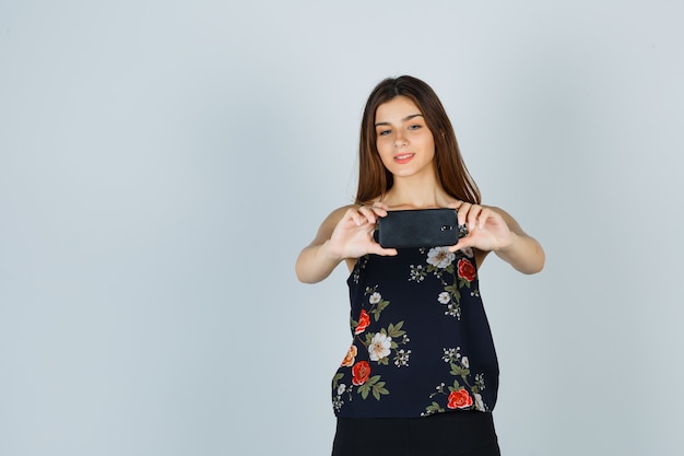 Young woman taking photo on smartphone in blouse and looking cheery , front view.