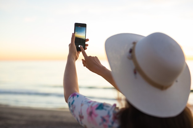 휴대폰이나 스마트폰 디지털 카메라로 바다와 일몰 사진을 찍는 젊은 여성