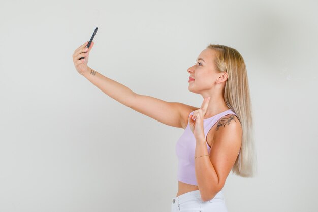 Young woman taking selfie by showing v-sign in singlet, mini skirt .