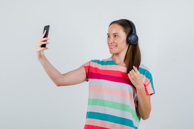 Young woman taking selfie on smartphone in t-shirt, headphones and looking cheerful. front view.