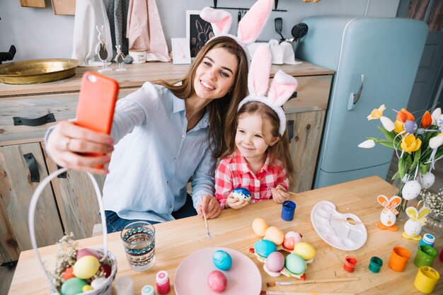 Young woman taking selfie with daughter near Easter eggs 